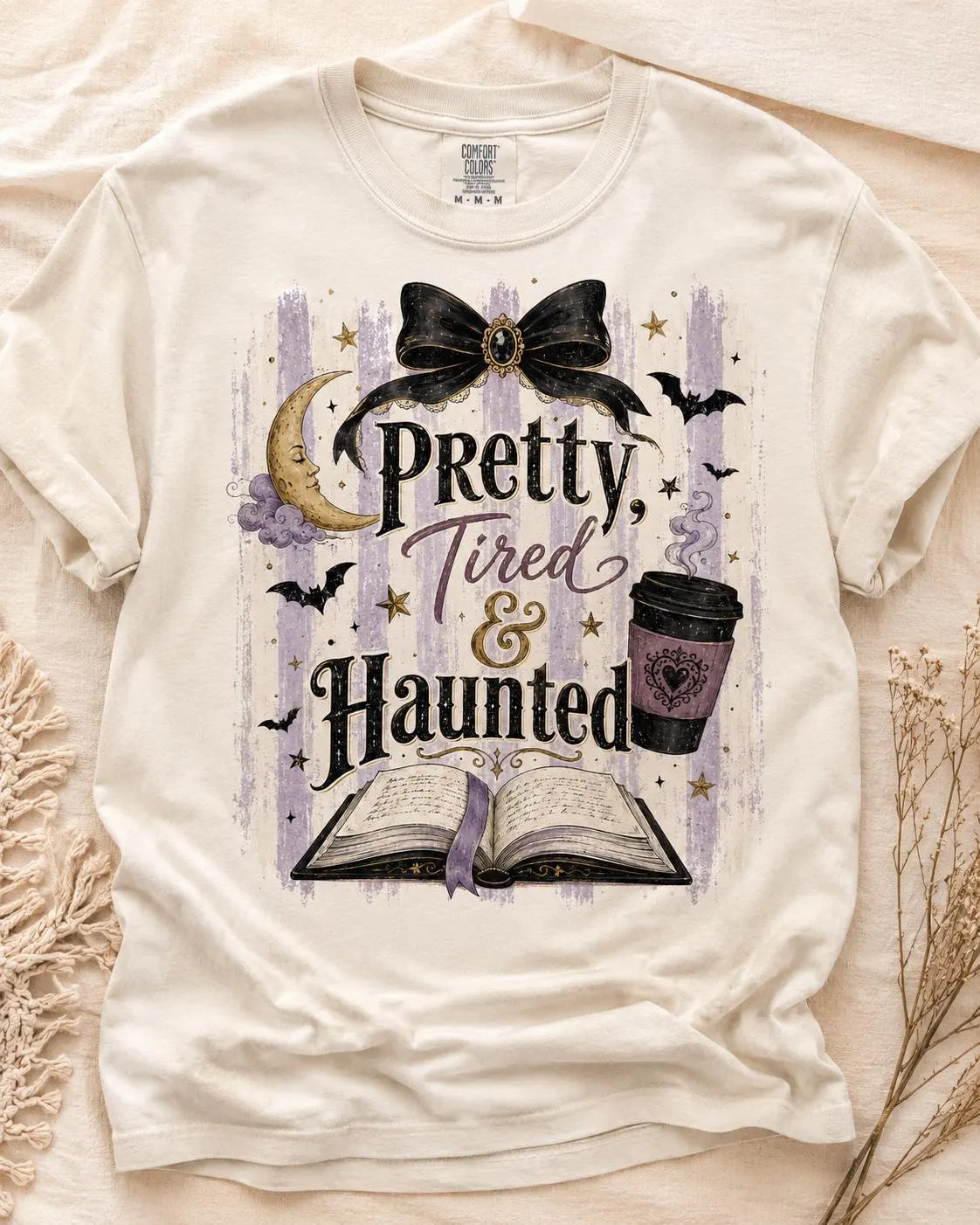 Preview of Pretty, Tired & Haunted lettering, an open book, coffee, a black bow, a crescent moon and bats on a shirt mockup